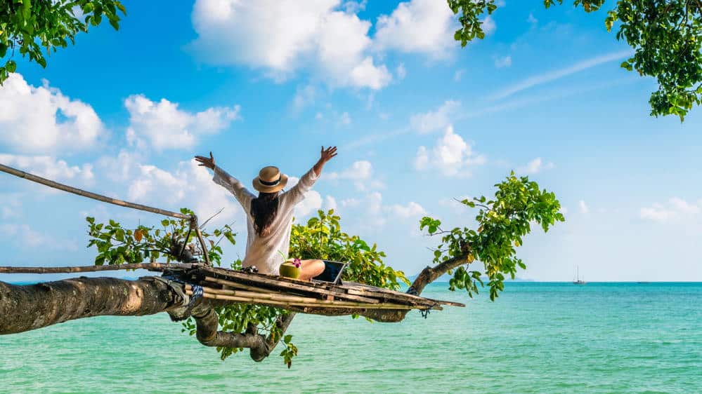 a woman relaxes on a phuket beach sitting on a branch with her laptop placed in her lap and her hands extended